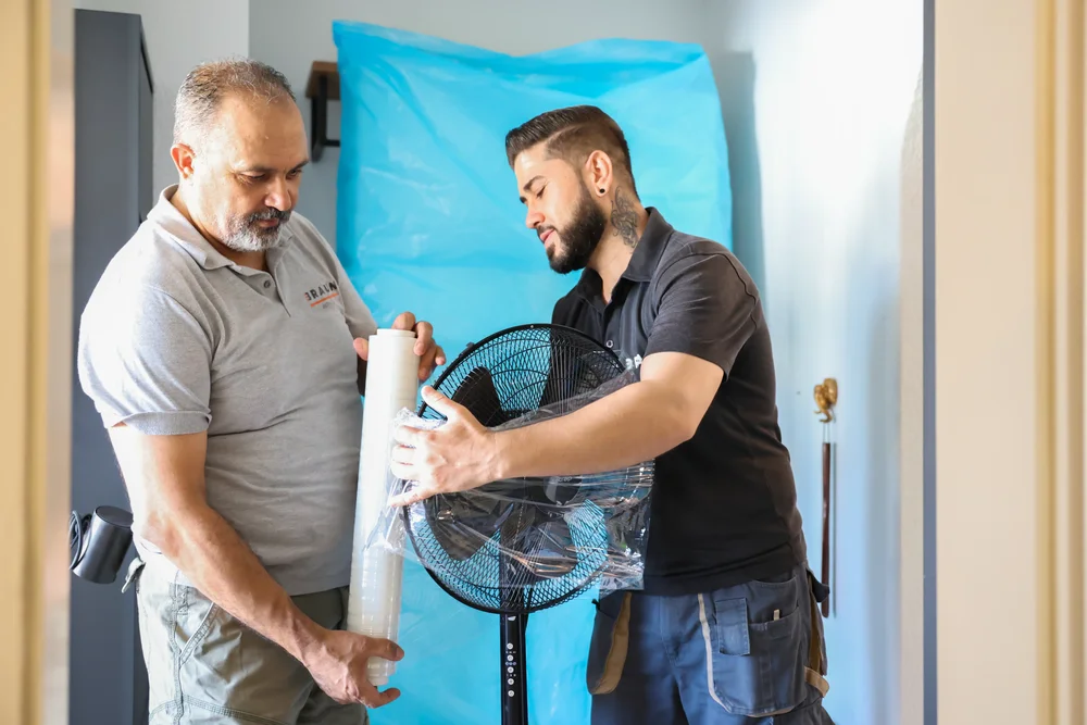 Zwei Mitarbeiter bereiten einen Ventilator mit Schutzmaterial für den Transport vor.